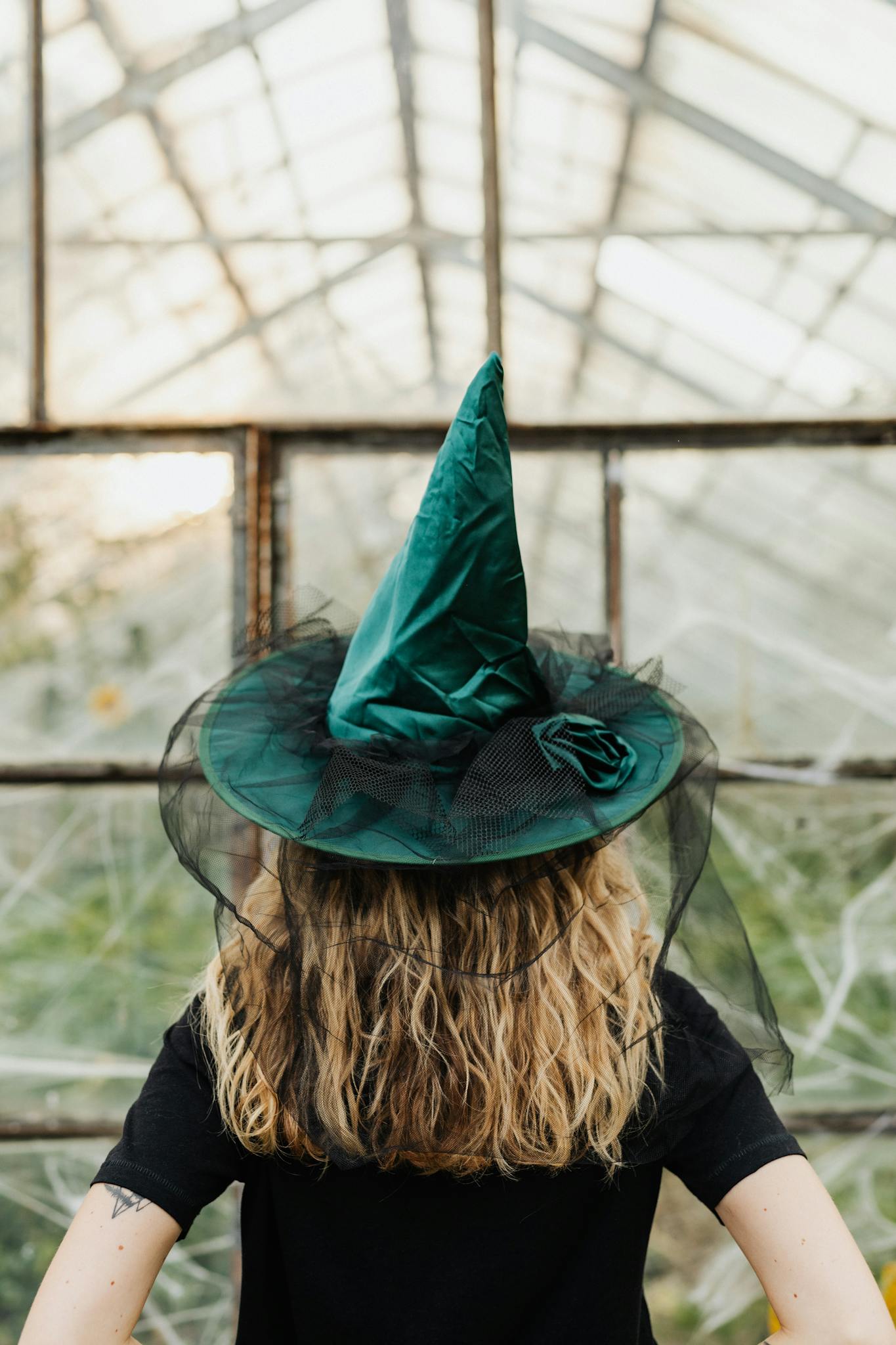 A woman with long hair wearing a green witch hat in a greenhouse setting.